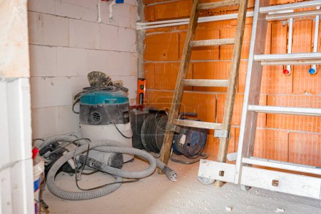 Wooden and metal ladders leaning against wall made of red blocks, placed in corner of room under construction, with industrial vacuum cleaner and few electric extension cords with reel.