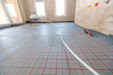 Long thin white tube unrolled over divided surface, with red auxiliary lines, grid, for help in placing pipeline of underfloor heating system in a residential building, brass for central heating floor in home.