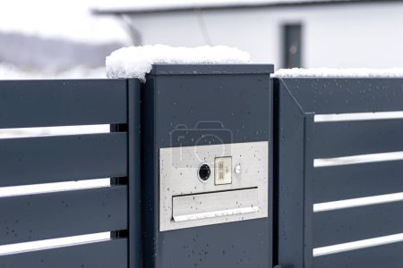 Téléchargez les photos : A modern videophone with a letterbox and a wireless card reader, mounted in panel fence in anthracite color, visible wicket, it rains in winter. - en image libre de droit