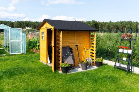 Foto de Un cobertizo de jardín de madera de pie sobre una base de hormigón en un jardín, flores y herramientas visibles. - Imagen libre de derechos