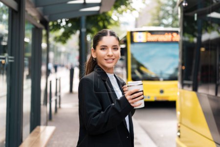 Téléchargez les photos : Belle jeune femme d'affaires hispanique multiraciale utilisant les transports en commun dans la ville souriant et tenant eco thermotasse avec café. - en image libre de droit