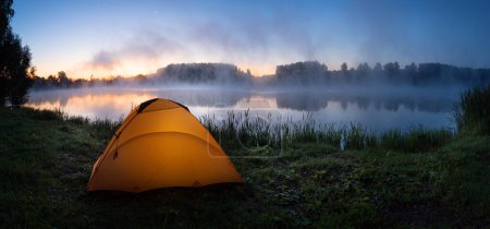 Photo for Orange tent lit from the inside on the bank of a foggy river in the - Royalty Free Image
