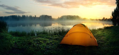 Photo for Orange tent on the bank of the river covered with fog in the early morning - Royalty Free Image