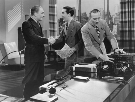 Foto de Felices colegas masculinos estrechando las manos cerca del hombre de negocios usando la radio en el escritorio en la oficina - Imagen libre de derechos