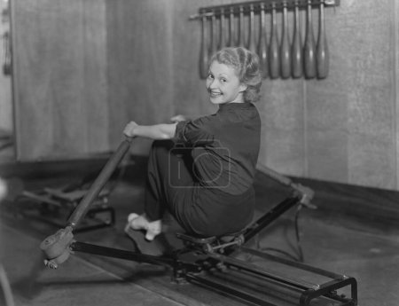 Foto de Mujer joven sonriente mirando por encima del hombro mientras hace ejercicio en el remo en el gimnasio - Imagen libre de derechos