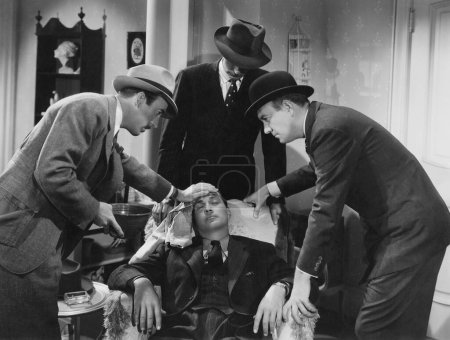 Foto de Amigos vistiendo sombreros cuidando a un hombre enfermo sentado inconsciente en una silla en casa - Imagen libre de derechos