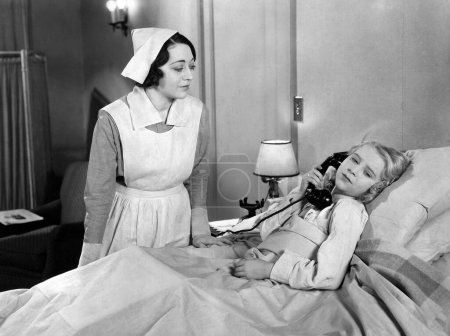 Foto de Enfermera mirando al paciente adolescente hablando a través del receptor telefónico mientras está acostado en la cama en el hospital - Imagen libre de derechos