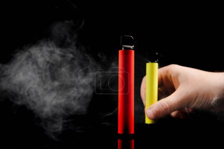 Disposable electronic cigarettes of different flavors in hand on a yellow background. The concept of modern smoking, vaping and nicotine