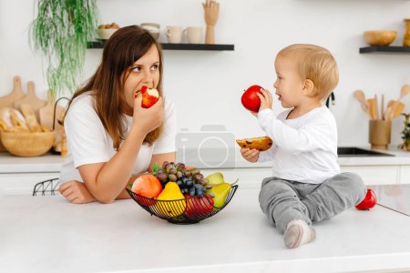 Photo for Mom and her son are in the kitchen eating healthy food. the baby is holding a cookie and an apple. Reducetarian Eating Enjoying Plant-Based Food. - Royalty Free Image