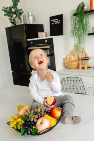 Foto de El niño come una manzana para desayunar sentado en la cocina. Alimentación saludable para toda la familia. Disfrutar de alimentos a base de plantas - Imagen libre de derechos