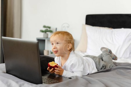 Photo for A smiling child lying on a bed next to his laptop. - Royalty Free Image