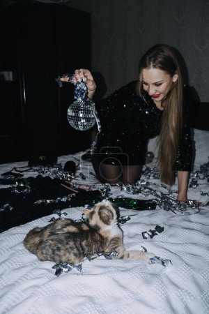 Photo for Nightlife, New Year Eve party. Beautiful woman in shiny evening dress is going to New Years Eve party. Young beautiful girl playing with the kitten mirror ball at home. - Royalty Free Image