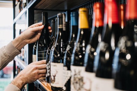 Cliente Seleccionando Botella de Vino del Estante de la Tienda. Una mano de las personas recogiendo una botella de vino de una selección diversa en un estante bien surtido de la tienda de vino.