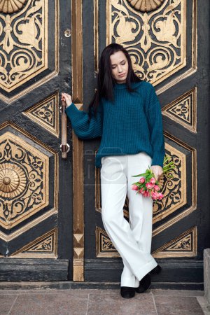 Seasonal Changes, Spring Fashion. A thoughtful woman in a chic blue sweater and white pants holds pink tulips while leaning on a beautifully carved antique door, showcasing spring fashion.