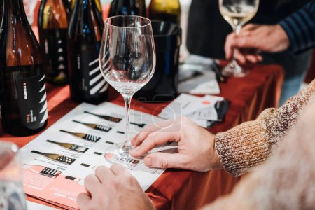 Téléchargez les photos : Un participant examine un verre de vin sur une feuille de dégustation lors d'une dégustation professionnelle de vin, avec une variété de bouteilles en arrière-plan. - en image libre de droit