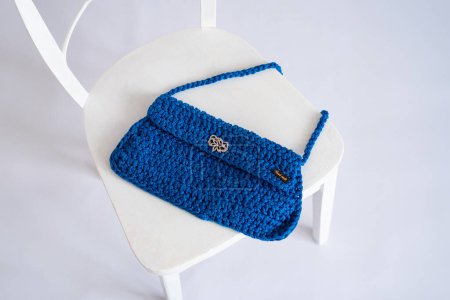 A vibrant blue crocheted clutch featuring a delicate floral metal clasp, displayed on a white chair, highlighting eco-friendly handcrafted designs.