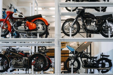 Foto de Un diverso conjunto de motocicletas clásicas, cada una con diseños y colores únicos, exhibidas en estanterías de metal industrial en un entorno de museo. - Imagen libre de derechos