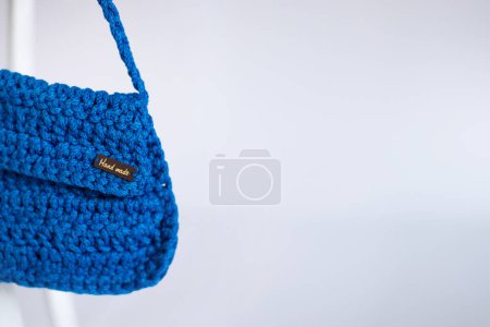 Embrayage au crochet bleu vif avec texte sans marque Fait à la main sur l'étiquette, affiché sur une chaise blanche, mettant en évidence des designs artisanaux respectueux de l'environnement.