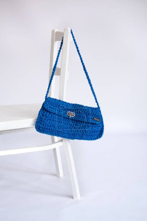 A vibrant blue crocheted clutch with unbranded text Handmade on the tag, displayed on a white chair, highlighting eco-friendly handcrafted designs.