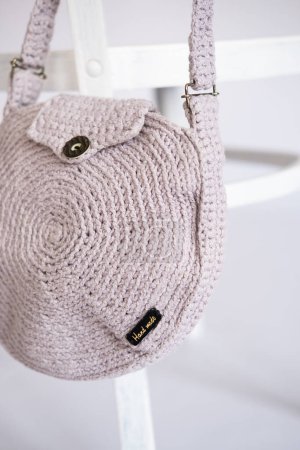 crafted handmade crocheted shoulder bag with unbranded text Handmade on the tag in a soft pink hue, displayed on a white chair, symbolizing artisanal fashion and creativity.