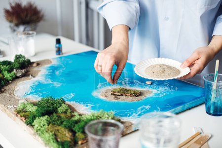 An artist meticulously adds sand details to her 3D epoxy resin artwork, creating a realistic coastal scene, demonstrating intricate artistic techniques in her bright studio.
