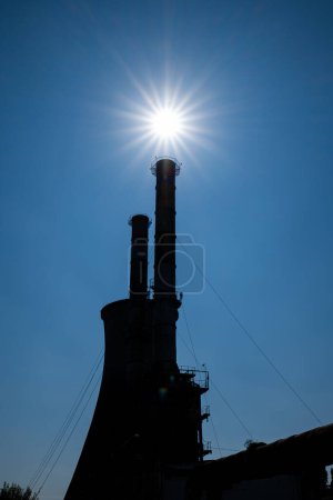 Téléchargez les photos : Cheminée industrielle ou cheminées de fumée silhouette face au soleil d'été de midi. Modèle de soleil éclatant d'étoiles, ciel bleu clair, personne. - en image libre de droit