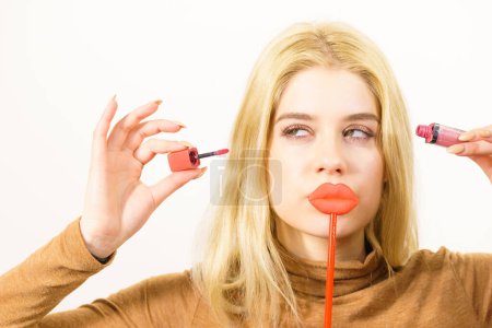 Junge erwachsene Frau, die Lippenstift oder Lipgloss aufträgt und sich schminkt, indem sie falsche Lippen am Stock hält