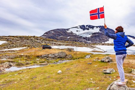 Mujer sostiene bandera noruega en las montañas, furgoneta con canoa en el techo superior en la distancia. Ruta turística nacional 55 Sognefjellet, Noruega