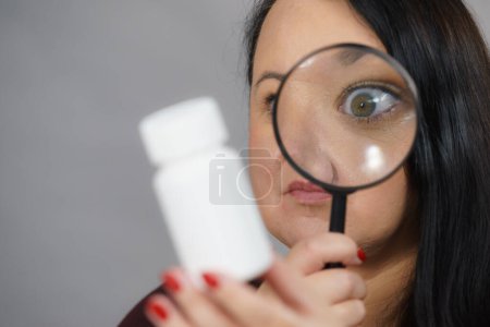 Mujer adulta investigando ingredientes de medicamentos, productos químicos utilizados en cápsulas píldoras dentro de la caja blanca. Hembra usando lupa