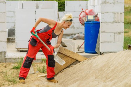 Woman working hard on construction site, using shovel digging sand soil. Partially built new home early stage. Industry.