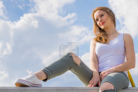Trendy fashionable woman relaxing outdoor wearing casual footwear white sneakers and hole trousers. Female enjoy sunlight against blue sky.