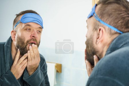 Hombre adulto divertido con máscara en la frente con problemas para despertarse. De pie frente al espejo mirando su cara, bostezando, cansado y soñoliento.