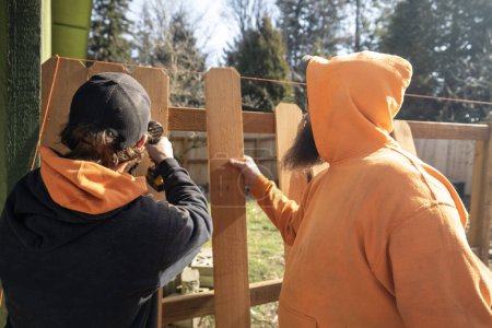 Téléchargez les photos : Deux travailleurs de la construction construisent une clôture extérieure Un homme aide un autre à aligner les lattes de piquet de bois sur un angle de niveau fixé par des vis avec outil électrique - en image libre de droit