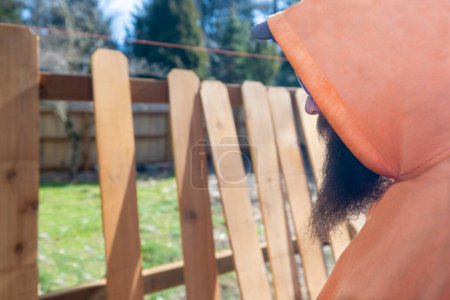 Photo for Fence Builder Construction Worker With Beard Examines and Measures Wood Pickets Along a Grass Yard - Royalty Free Image
