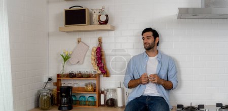 Photo for Handsome bearded man thinking while holding coffee cup in kitchen. Portrait of a young man looking at something through the kitchen window. - Royalty Free Image