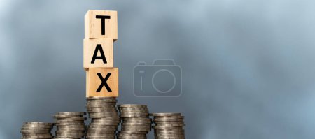 Tax concept with wooden blocks on stacked coins. Word TAX written on wooden cubes on stacked coins on dark background. Panoramic images have free space that can contain advertising media.