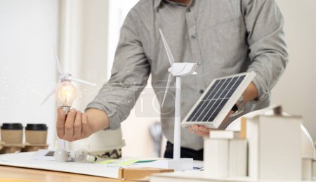 Foto de Close-up at a light bulb, An engineer sits holding a light bulb and model of wind turbine. with solar panels and model house on the table. To design use of renewable energy with wind and solar energy. - Imagen libre de derechos