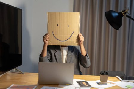 Un joven empresario sostiene un cartón con una cara sonriente delante de su cabeza, haciendo un gesto con una caja de cartón en su cabeza con una cara sonriente.