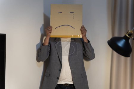 Foto de Un empleado infeliz con caja en lugar de su cabeza. Retrato del hombre de negocios cubriéndose la cara con una máscara de papel con la cara triste dibujada en ella, mala depresión negativa persona anónima. - Imagen libre de derechos