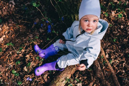 Téléchargez les photos : Petite fille en bottes violettes et globalement bleue assise sous l'arbre dans la forêt printanière couverte de fleurs violettes Hepatica nobilis - en image libre de droit