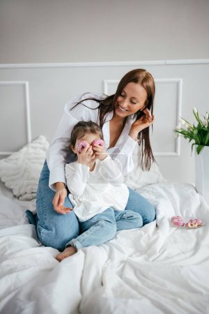 Photo for Happy young mom playing with her child, girl holding sweet doughnuts behind her eyes. Mother and daughter having fun together - Royalty Free Image