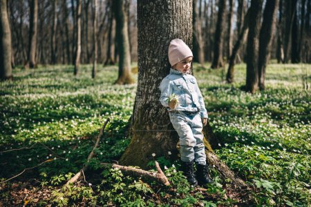 Retrato de adorable niña de 3 años en chaqueta vaquera de pie cerca del árbol en el bosque cubierto de anémonas sosteniendo ramo de flores del bosque. Feliz infancia. 
