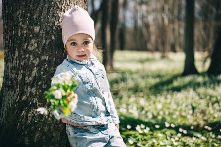 Portrait of adorable 3 years old girl in jeans jacket standing near the tree in the forest covered with anemones holding bouquet of forest flowers. Happy childhood 