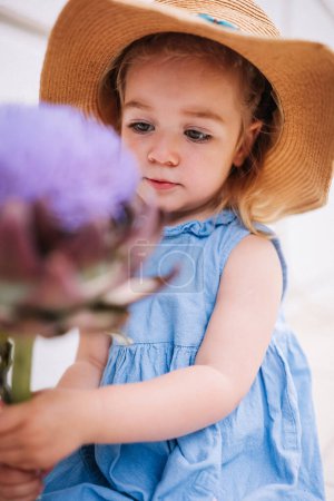 Portrait of cute little 2 years old girl in straw hat holding big artichoke flower. Happy childhood, summer activities