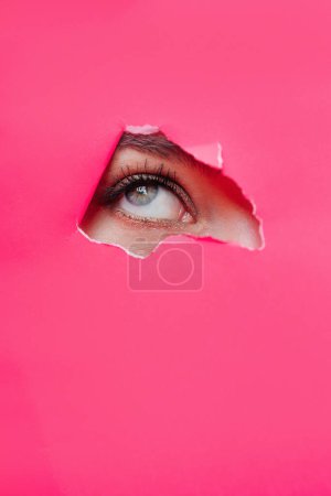 Téléchargez les photos : Œil de la belle femme regardant dans le trou en papier rose. Glamour femme beauté - en image libre de droit