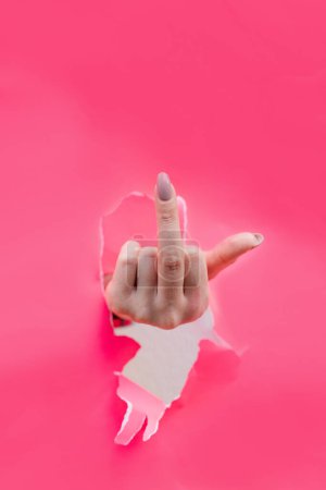 Téléchargez les photos : Femme main montrant majeur à travers un trou déchiré dans du papier rose, geste inapproprié. - en image libre de droit
