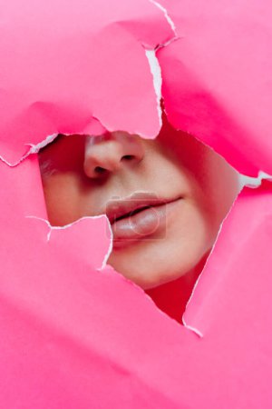 View of beautiful young woman with perfect lips through hole in pink paper. Plumped lips with injection fillers, woman beauty procedures