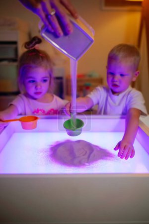 Foto de Adorable niño y niña de 3 años jugando en caja sensorial con tamices de arena y arena en la habitación de los niños. - Imagen libre de derechos