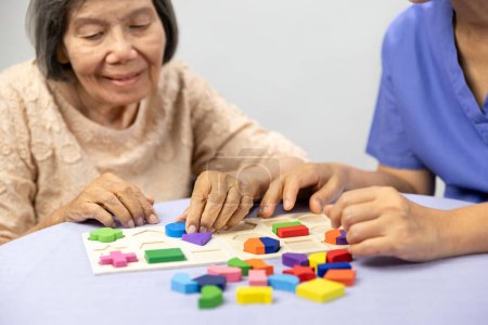 Téléchargez les photos : Aide-soignant et femme âgée jouant jeu de puzzles en bois pour la prévention de la démence - en image libre de droit
