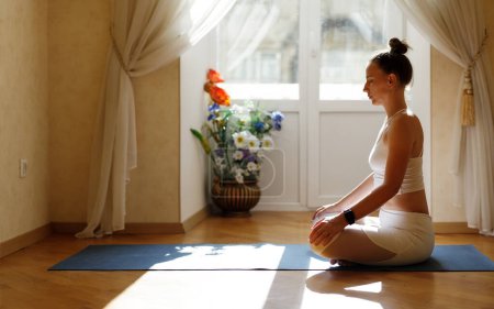 Fitness woman in white sportswear meditating on the mat near the balcony in the morning.